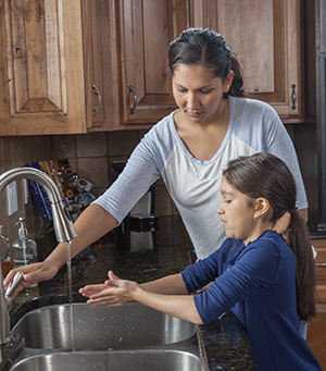 Woman helping girl wash hands in kitchen sink.