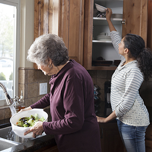 Una mujer extiende el brazo para tomar un objeto del estante superior del armario de la cocina mientras una mujer mayor prepara una ensalada.