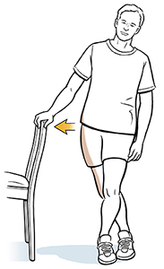 Un hombre sostiene una silla con una mano, con una pierna cruzada detrás de la otra. La flecha indica cómo inclina la cadera hacia la silla para estirar la banda iliotibial.